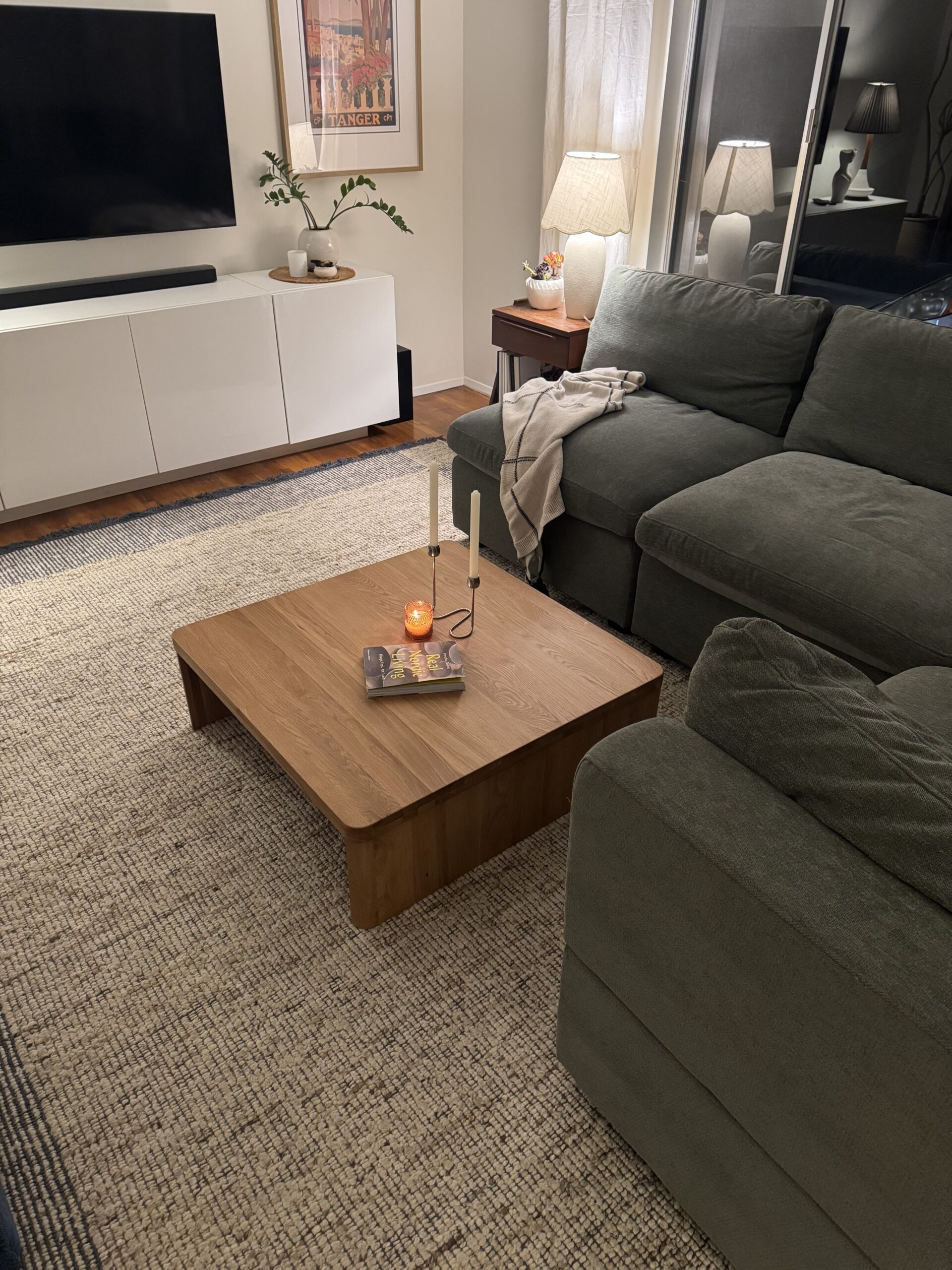 A living room with a green sectional sofa, wooden coffee table with candles, and a TV on a white media console. A floor lamp and framed art are in the background.