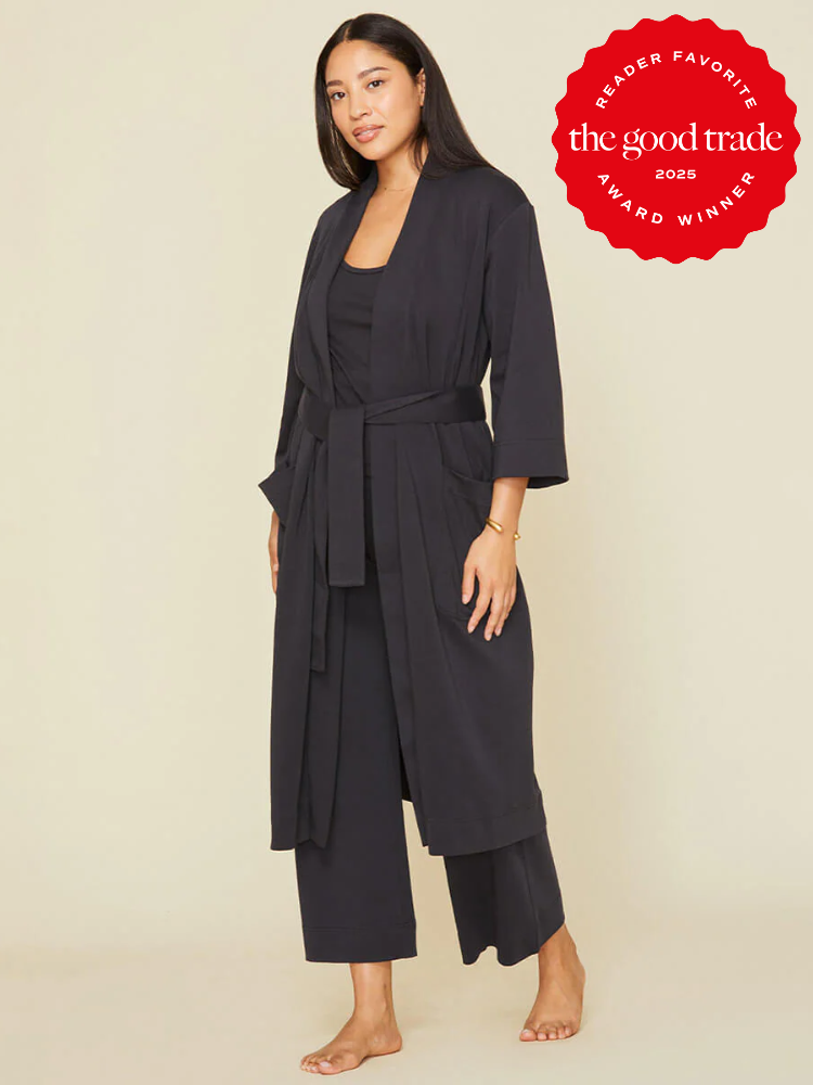 A woman stands barefoot wearing a black robe-style lounge set with wide sleeves and a waist tie; a red "The Good Trade Award Winner 2025" badge is in the upper right corner.