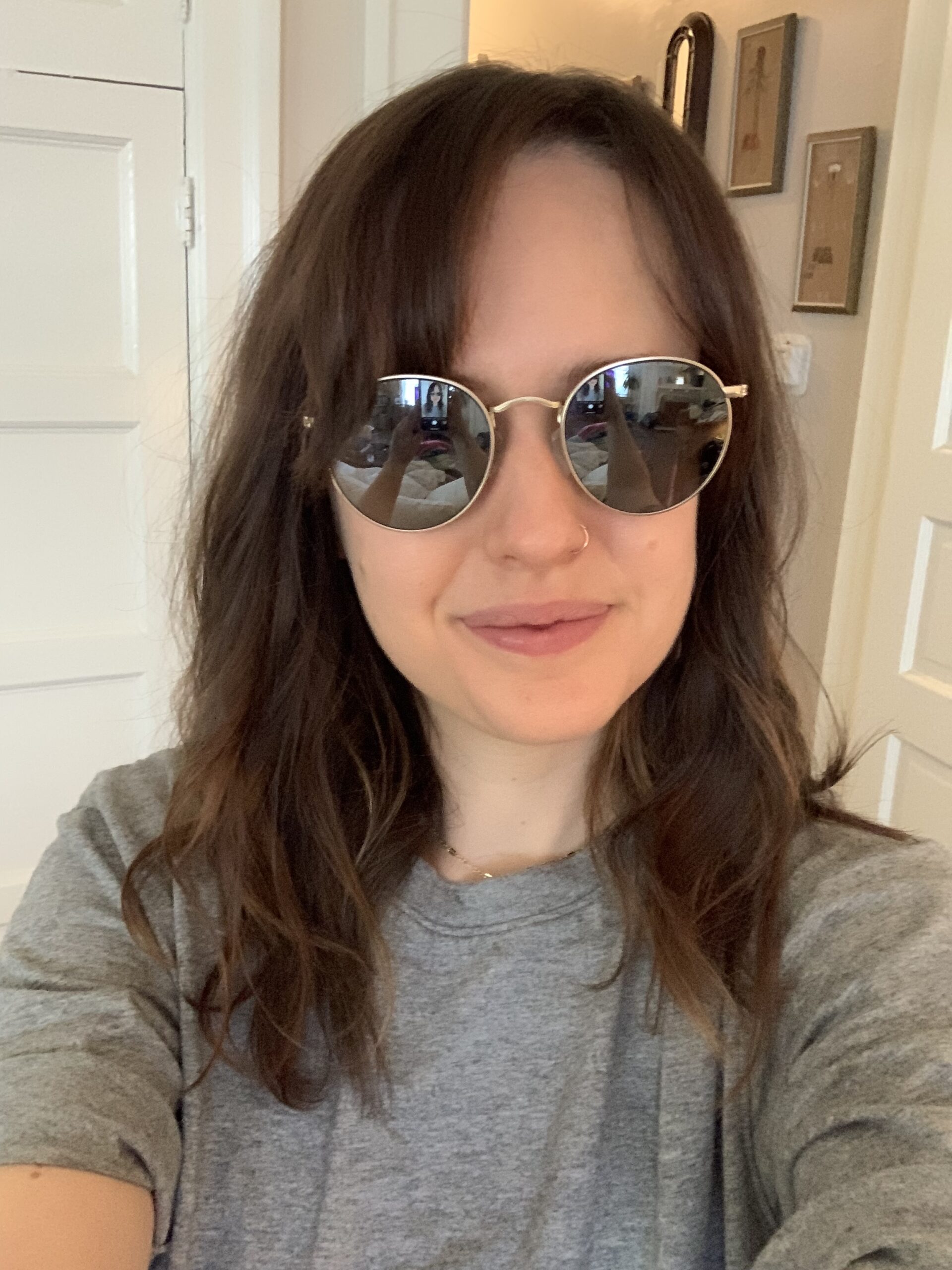 A woman with wavy brown hair wearing round sunglasses and a grey t-shirt stands indoors, smiling slightly at the camera.