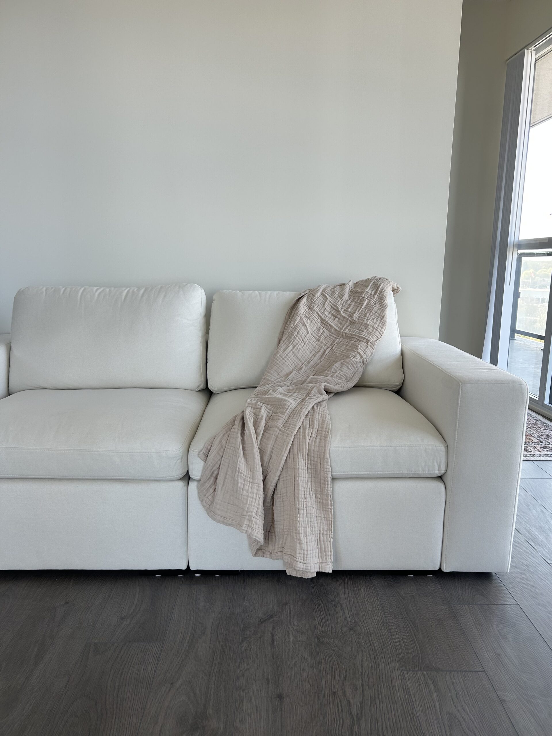 A beige blanket is draped over the arm and seat of a white sofa in a room with a light gray wall and dark wood flooring.