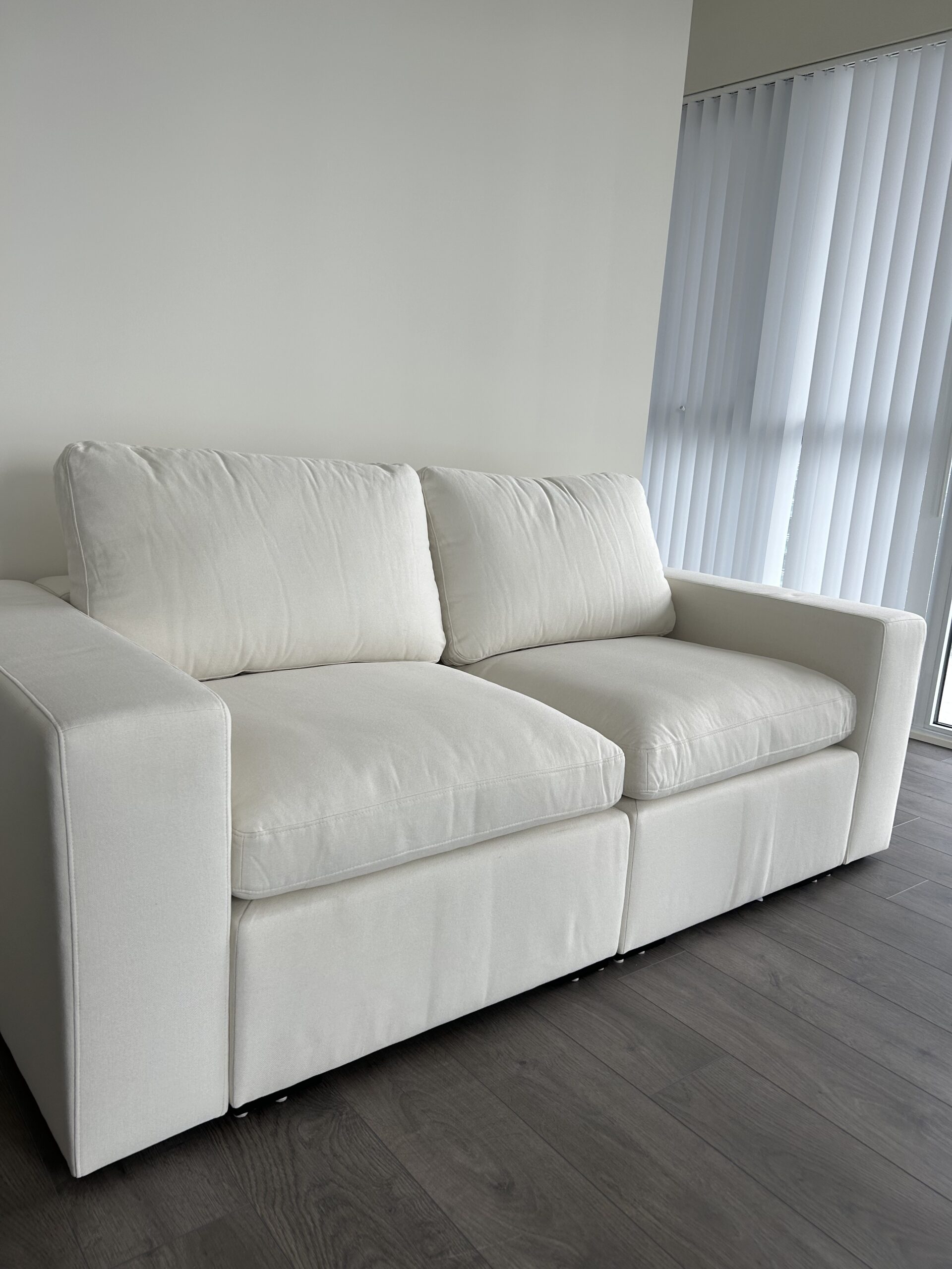 A white upholstered sofa with two seat cushions and two back cushions is placed against a light-colored wall on a dark wood floor near vertical window blinds.
