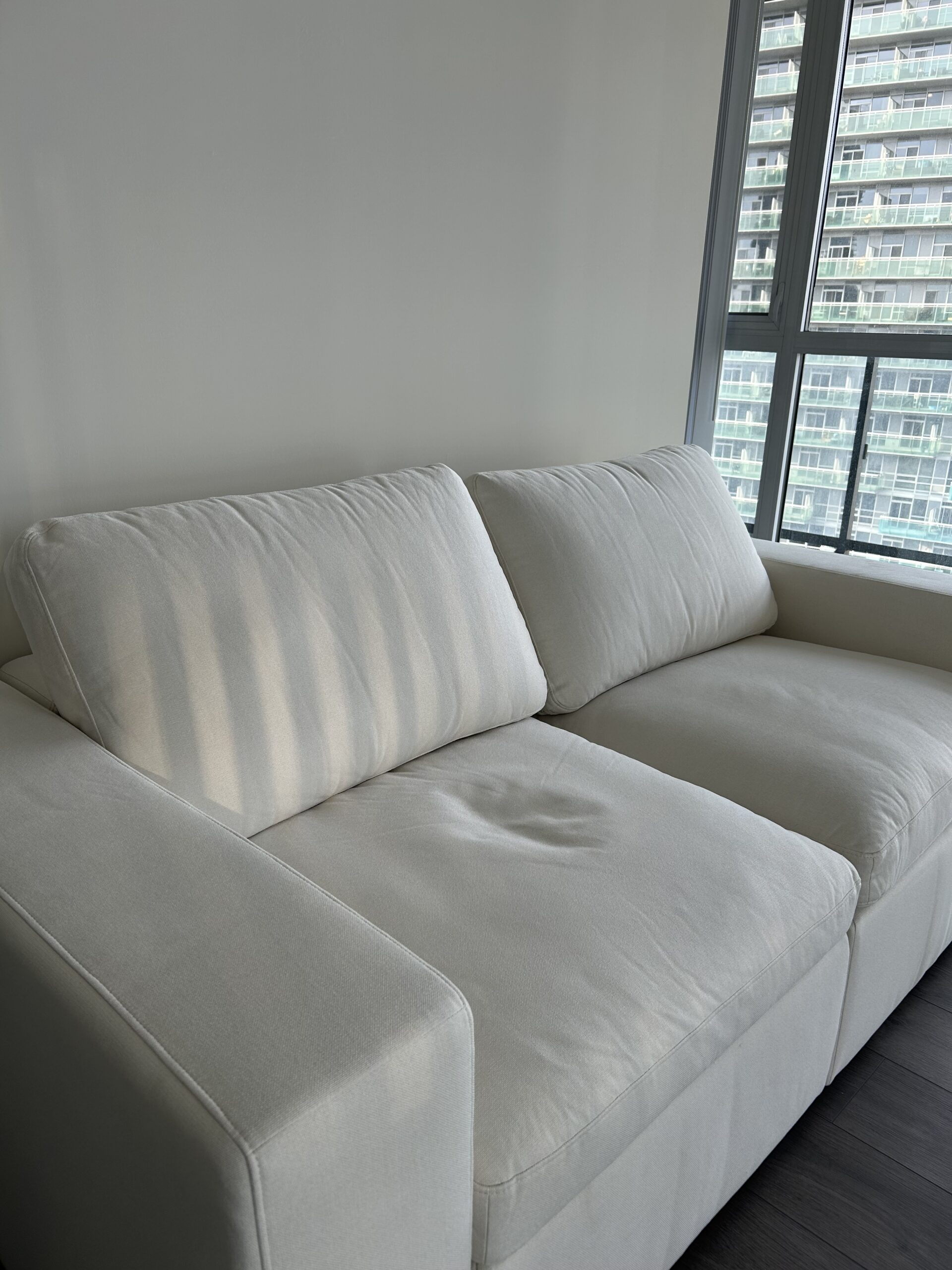 A white, modern two-seat sofa sits near a large window with vertical blinds, in a bright room with dark wooden floors.