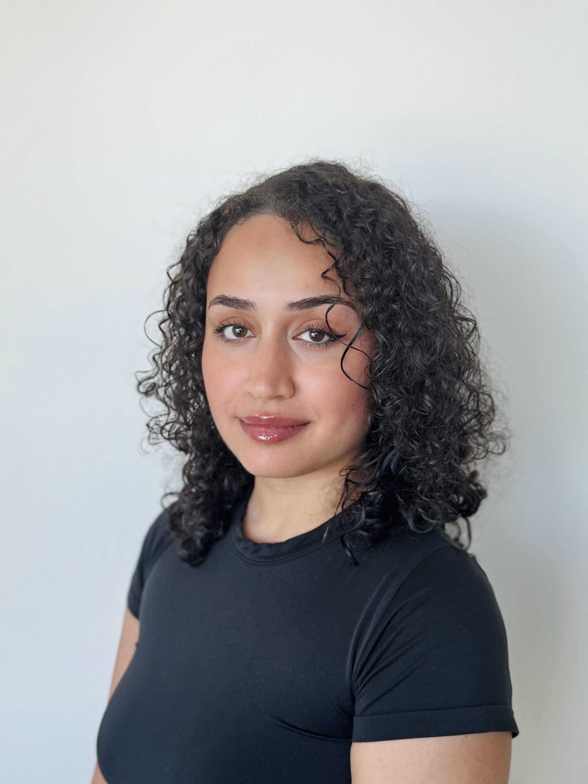 A person with medium skin tone and curly dark hair wears a black short-sleeved shirt, standing against a plain light background and looking at the camera.