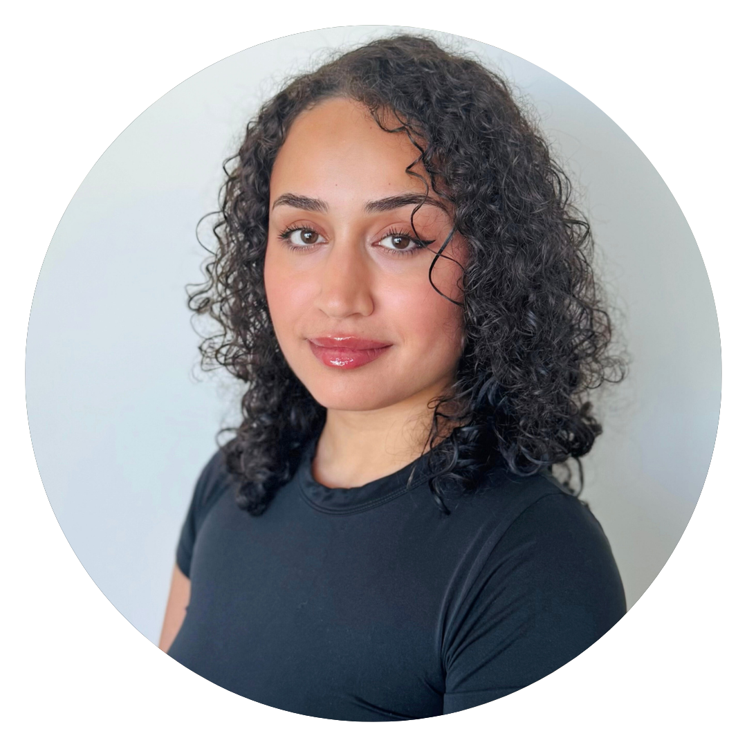 A woman with curly hair and a black t-shirt poses in front of a plain light-colored background, looking at the camera with a neutral expression.