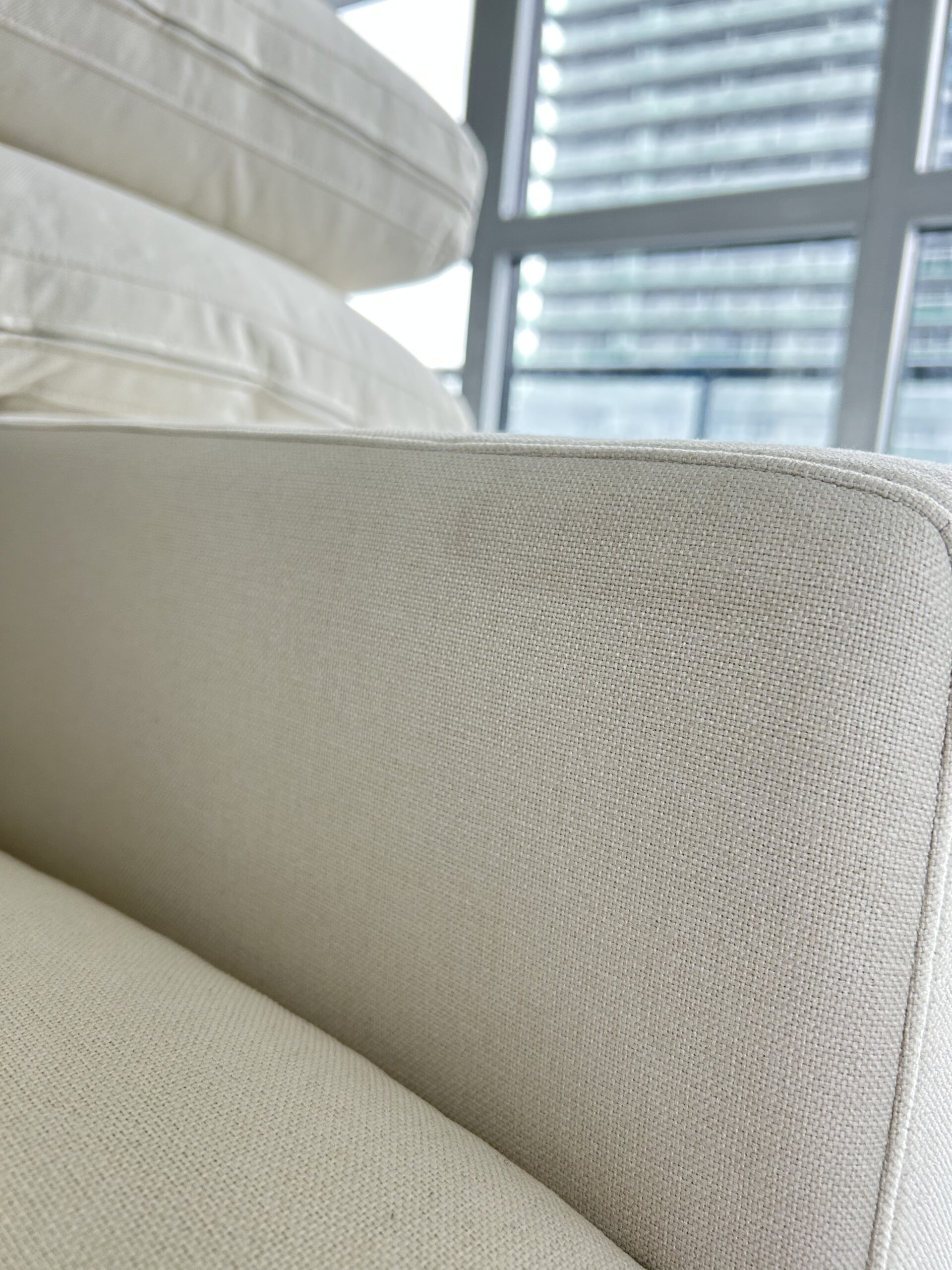 Close-up of a beige fabric sofa armrest with stacked cushions, set near large windows with a city building visible outside.