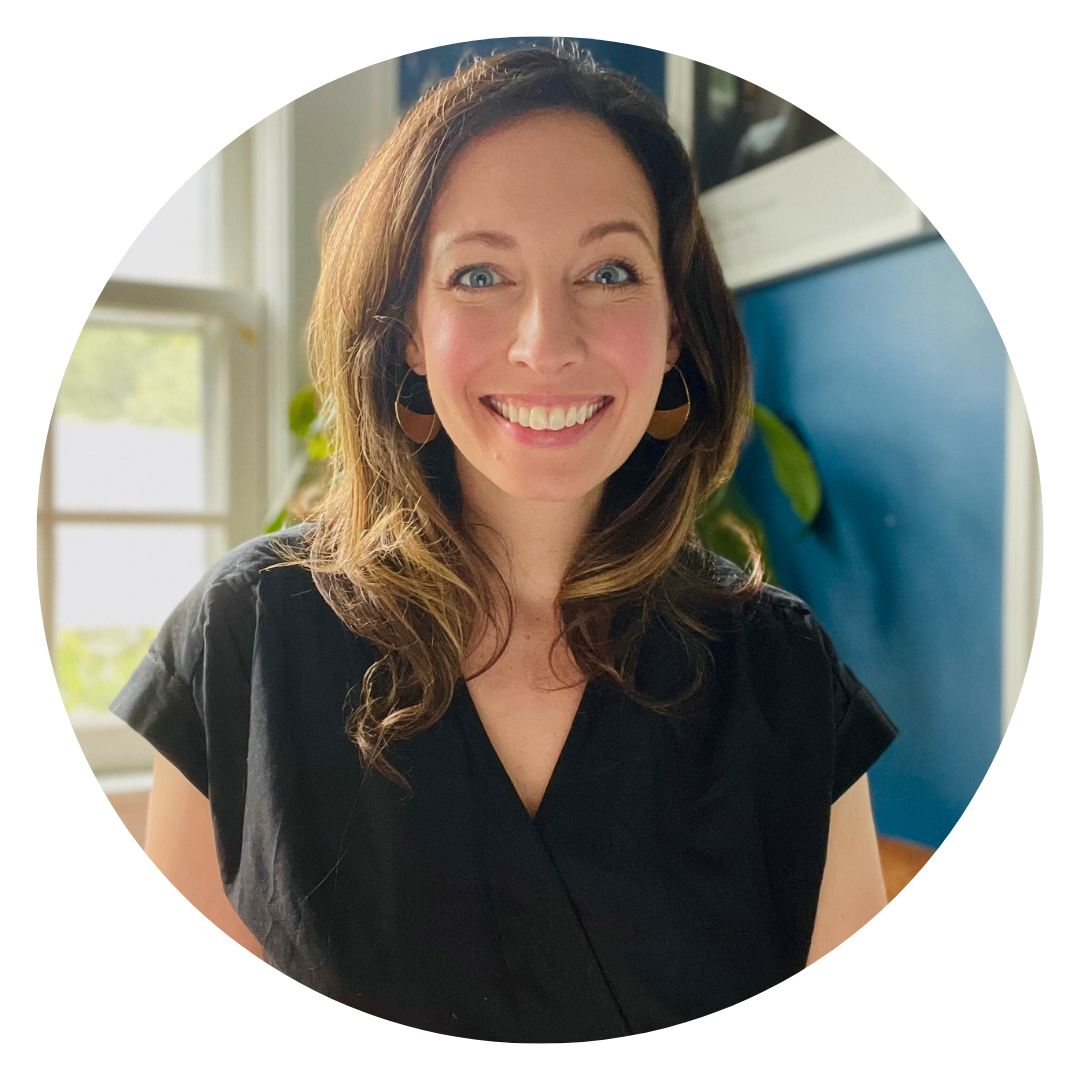 A woman with wavy brown hair and a black top smiles at the camera, standing indoors in front of a blue wall and a window.