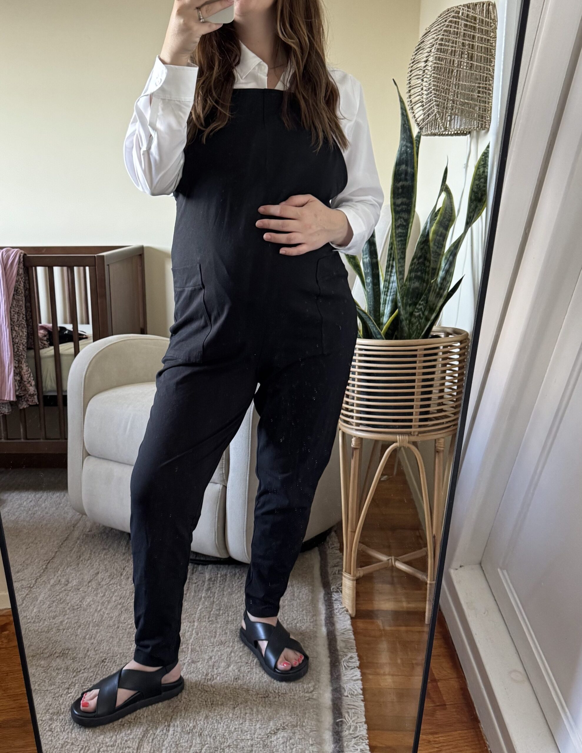 A woman takes a mirror selfie in a bedroom, wearing black overalls and sandals. There is a crib, a chair, a plant, and a lamp in the background.