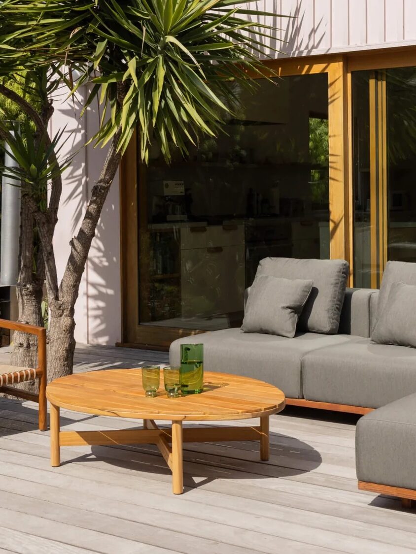 A modern outdoor patio with a gray sectional sofa, a wooden lounge chair, and a round coffee table on a wooden deck, next to a glass sliding door and tropical plants.