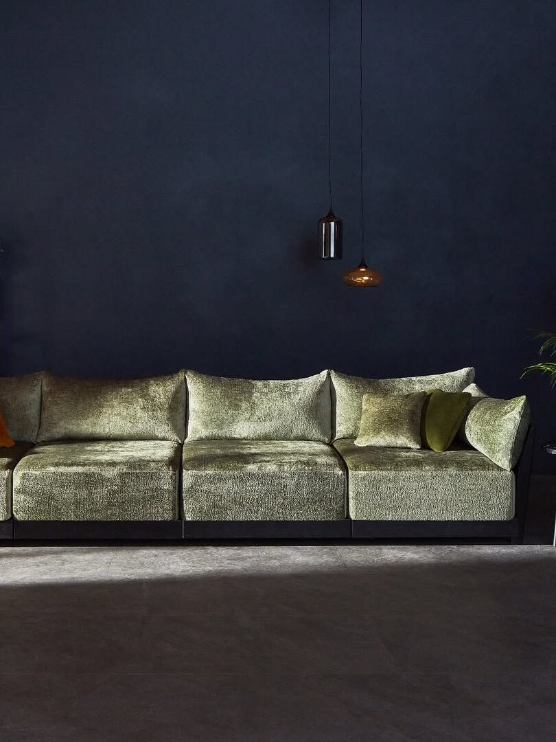 A large, light green velvet sectional sofa with several cushions sits against a dark blue wall, beneath two pendant lights. A bookshelf and potted plant are partially visible.