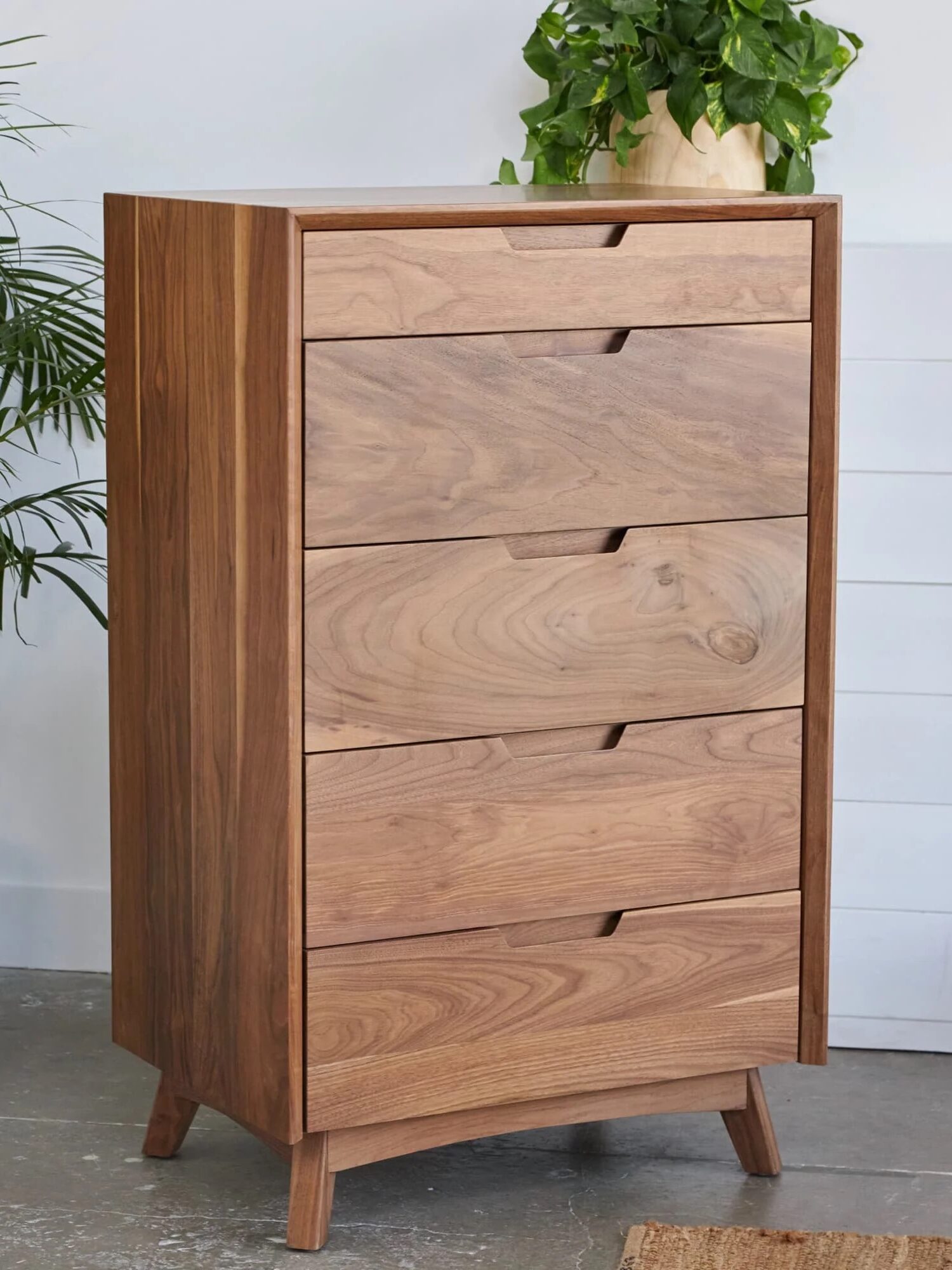 A tall, mid-century modern wooden dresser with five drawers stands on short angled legs, flanked by green potted plants in a bright room.
