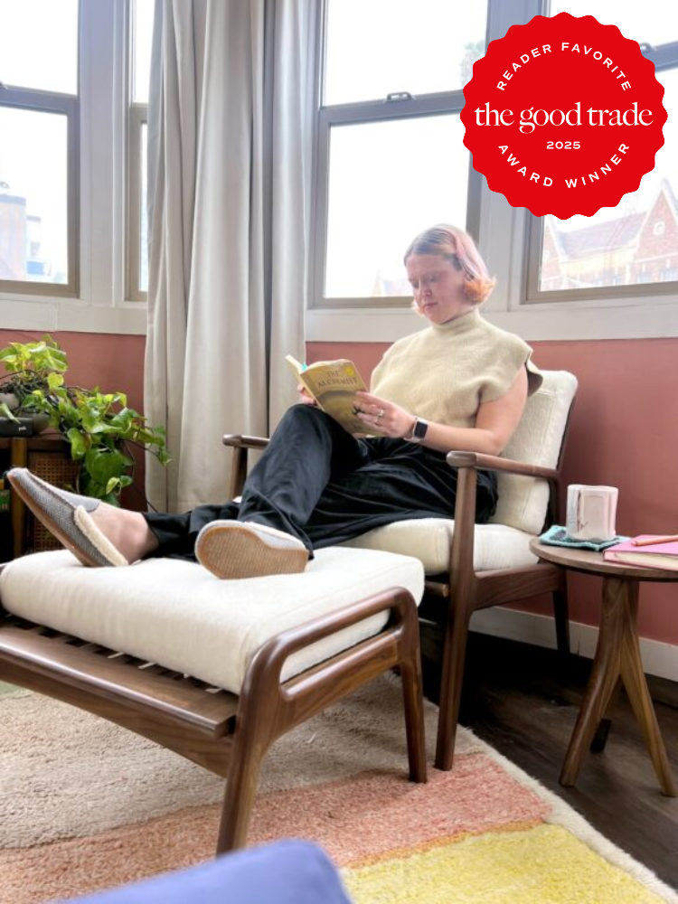 Person sitting on a wooden lounge chair reading a book in a sunlit room with plants, a side table, and large windows. A red award badge from The Good Trade is in the upper right corner.