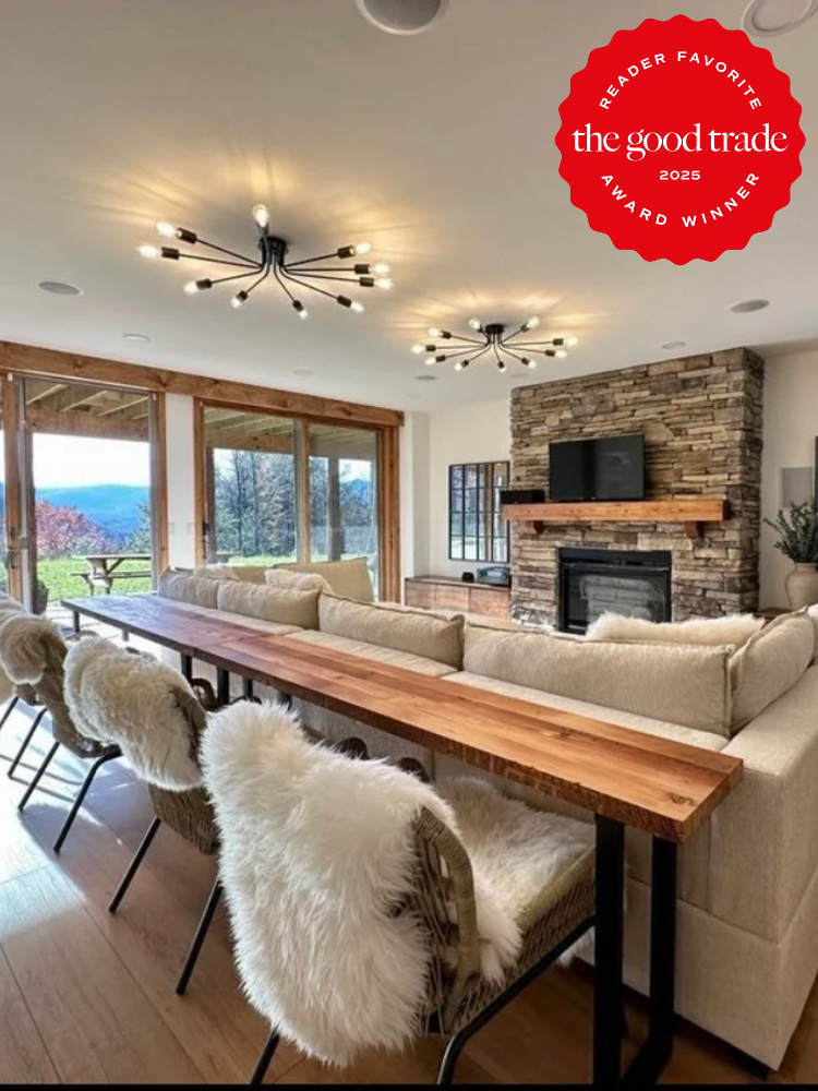 Modern living room with a stone fireplace, large windows, neutral furniture, fur-covered chairs at a wooden table, and contemporary ceiling lights. "The Good Trade 2025 Award Winner" badge in the corner.