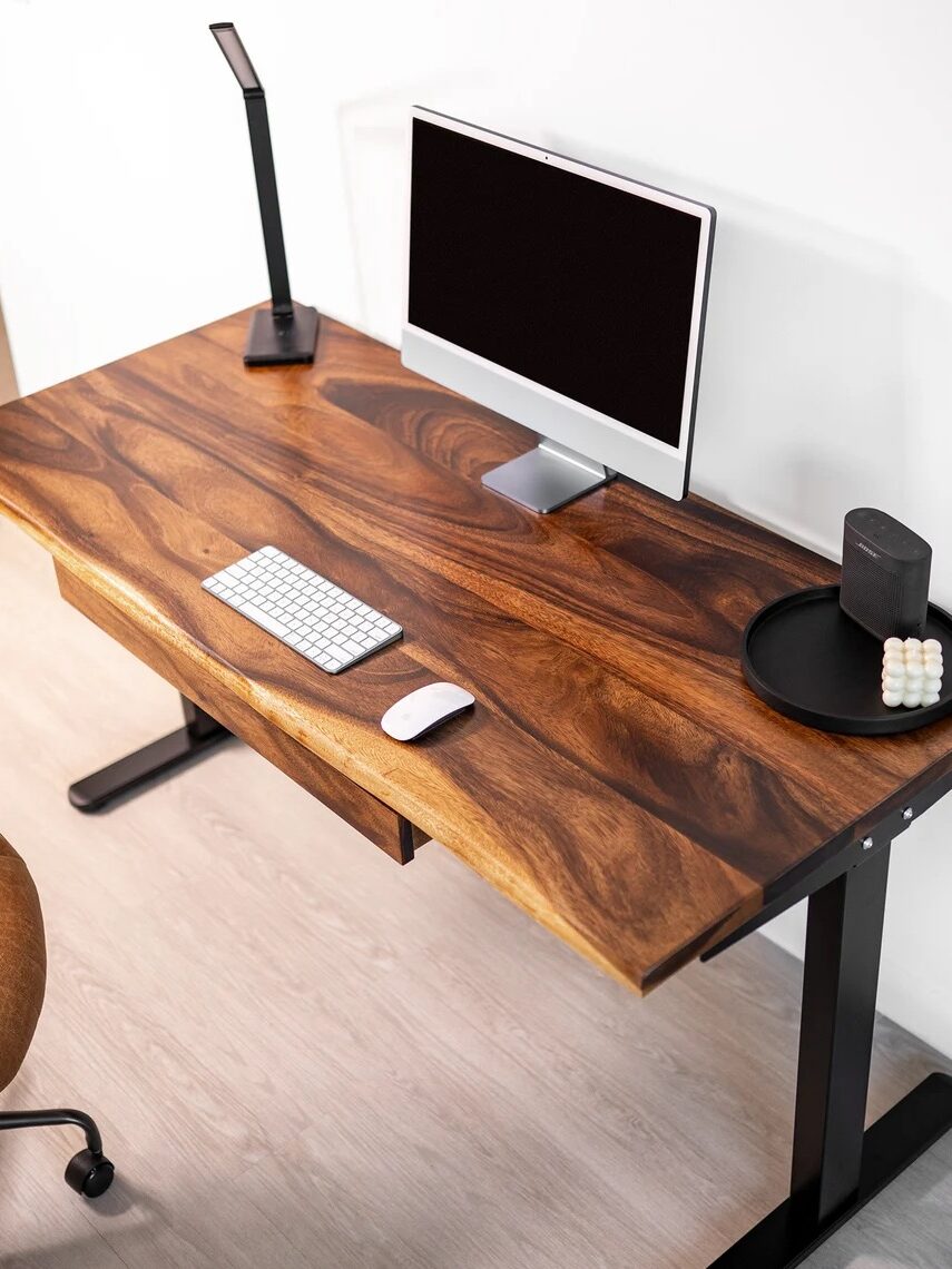 A wooden desk with a computer monitor, keyboard, mouse, desk lamp, speaker, and a tray with round objects, next to a brown chair on a light floor.