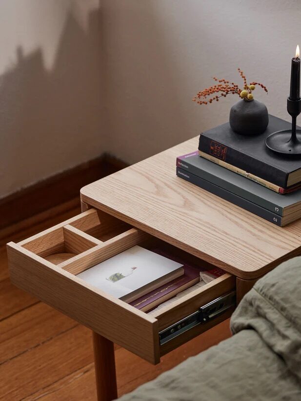 A wooden nightstand with an open drawer containing cards, and three stacked books, a vase, and a lit candle on top beside a bed with a green quilt.
