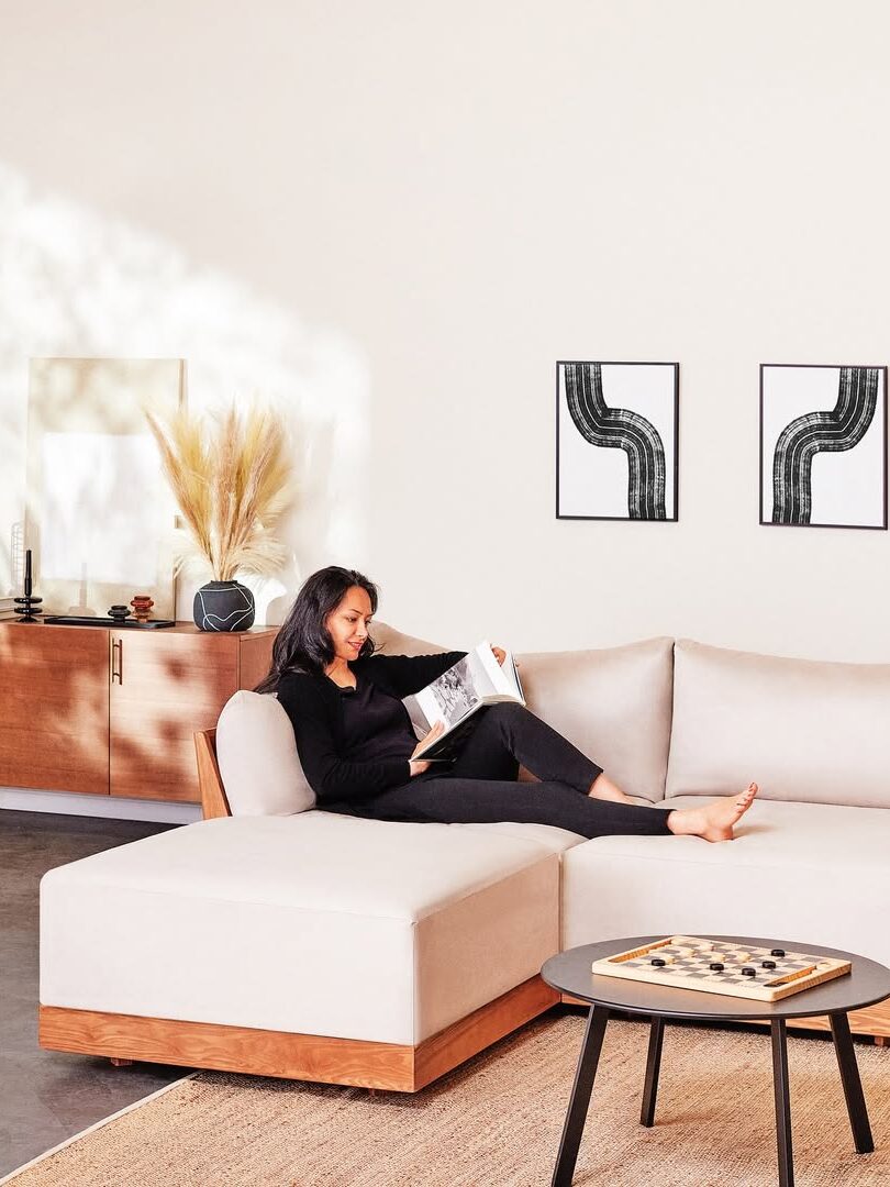A woman sits on a beige sectional sofa reading a magazine in a modern living room with minimalist decor and neutral tones.