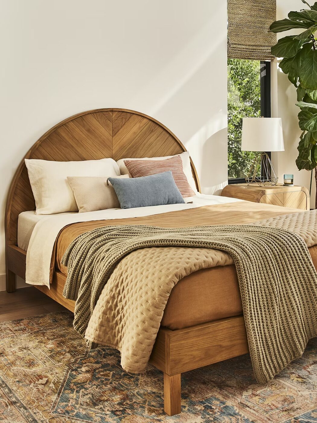 A neatly made bed with tan bedding and a wooden headboard sits between two matching nightstands, in a sunlit bedroom with large windows, plants, and a patterned rug.