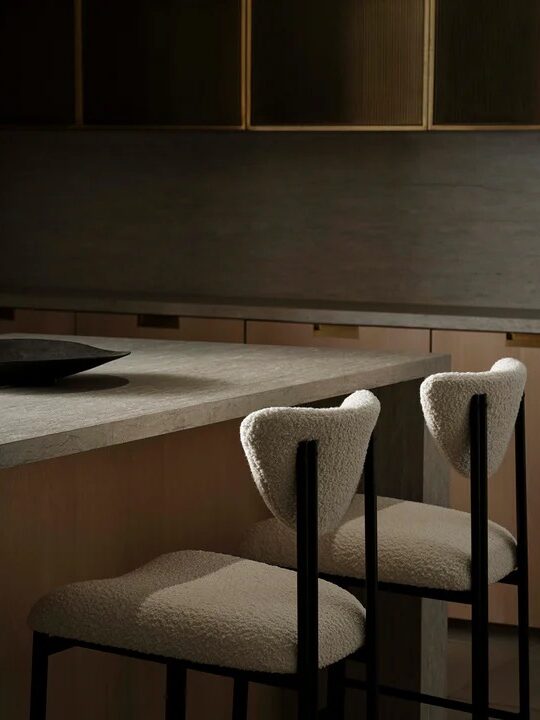Two modern barstools with textured white upholstery sit at a minimalist kitchen counter in a dimly lit room with wood cabinets.