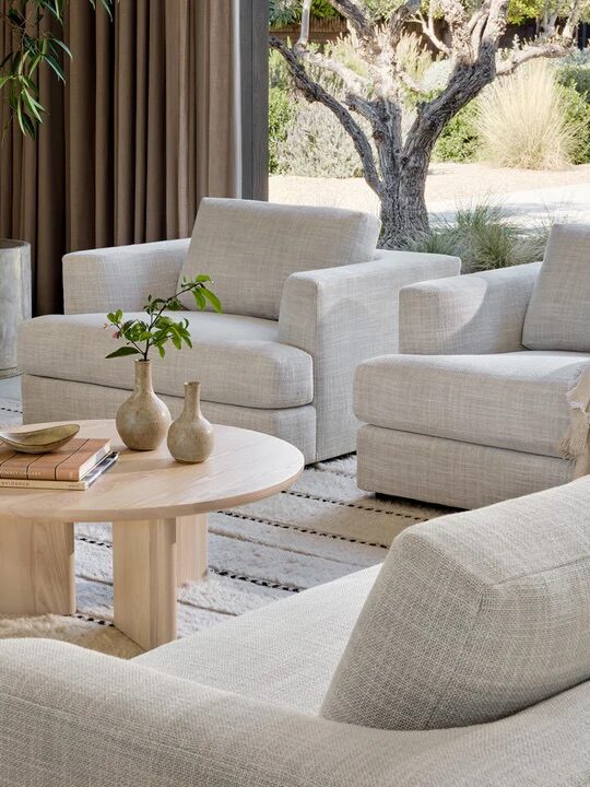A modern living room with light-colored sofas, a round wooden coffee table with vases and books, and large windows overlooking a garden.