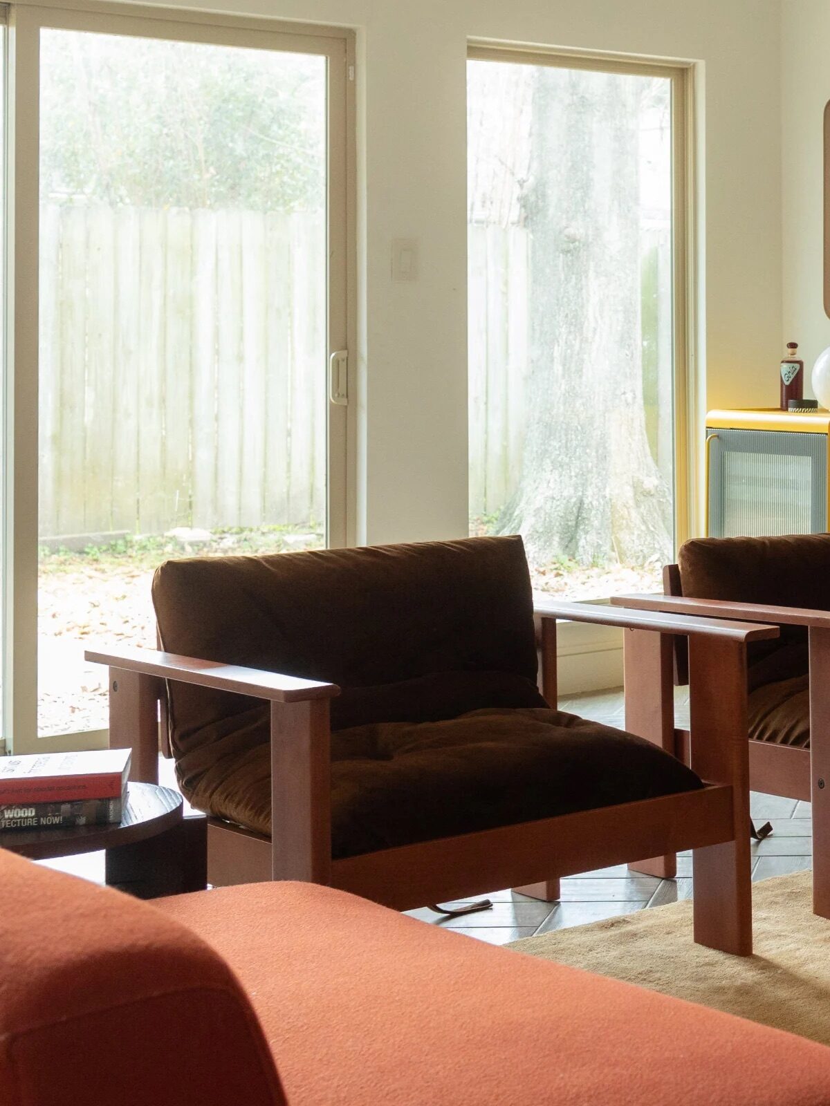 A living room with two brown armchairs, a red sofa, a beige rug, a stack of books, a framed poster, and large windows letting in natural light.