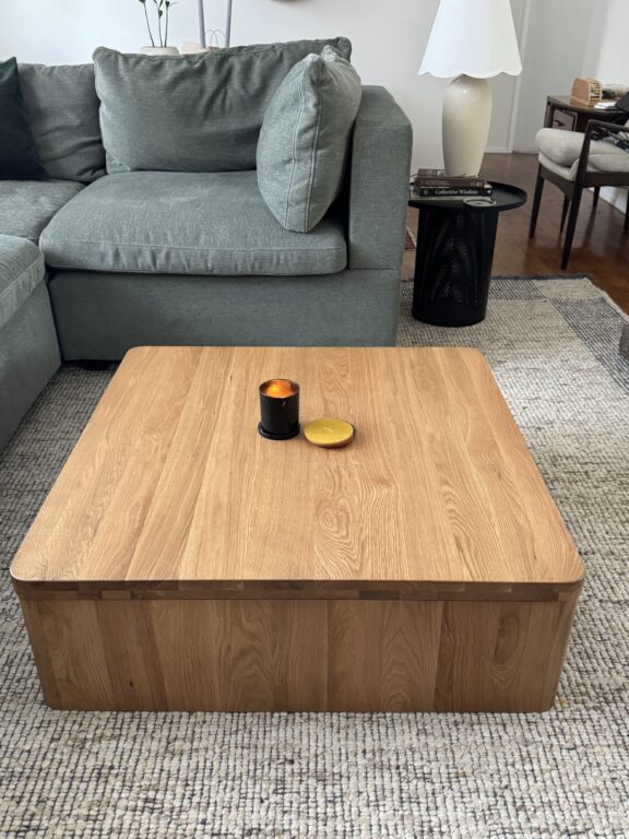A wooden square coffee table with a candle and a golden coaster sits on a textured rug in front of a gray sectional sofa in a modern living room.
