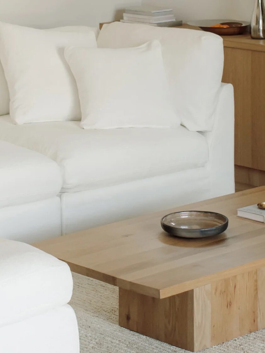 A minimalist living room with a white sectional sofa, a light wood coffee table holding a dish and a book, a beige rug, and a wooden sideboard with decor items.
