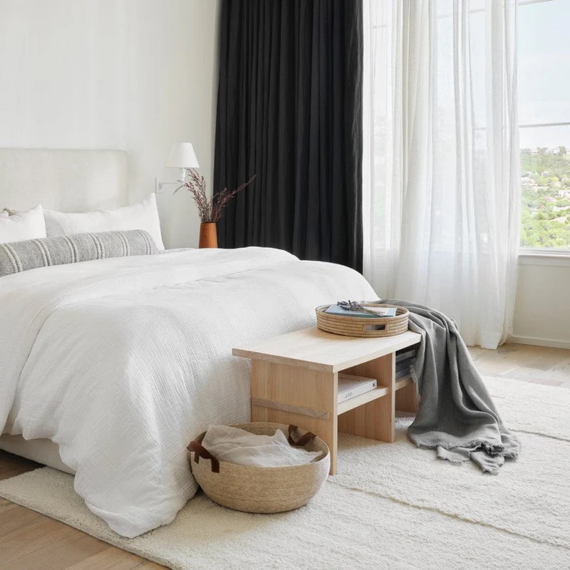 A modern bedroom with a neatly made bed, light wood bench, woven basket, gray throw, white rug, and large window with sheer and dark curtains.