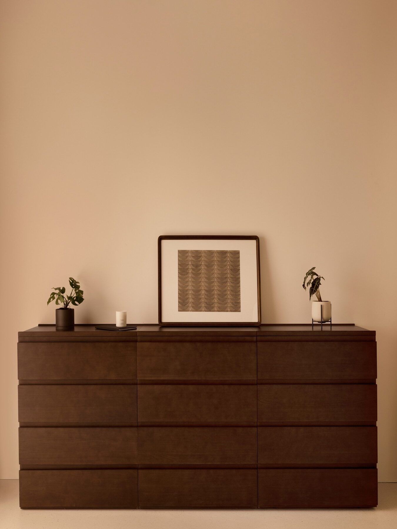 A dark wood dresser with a framed print, two small potted plants, and a candle arranged on top, set against a plain beige wall.