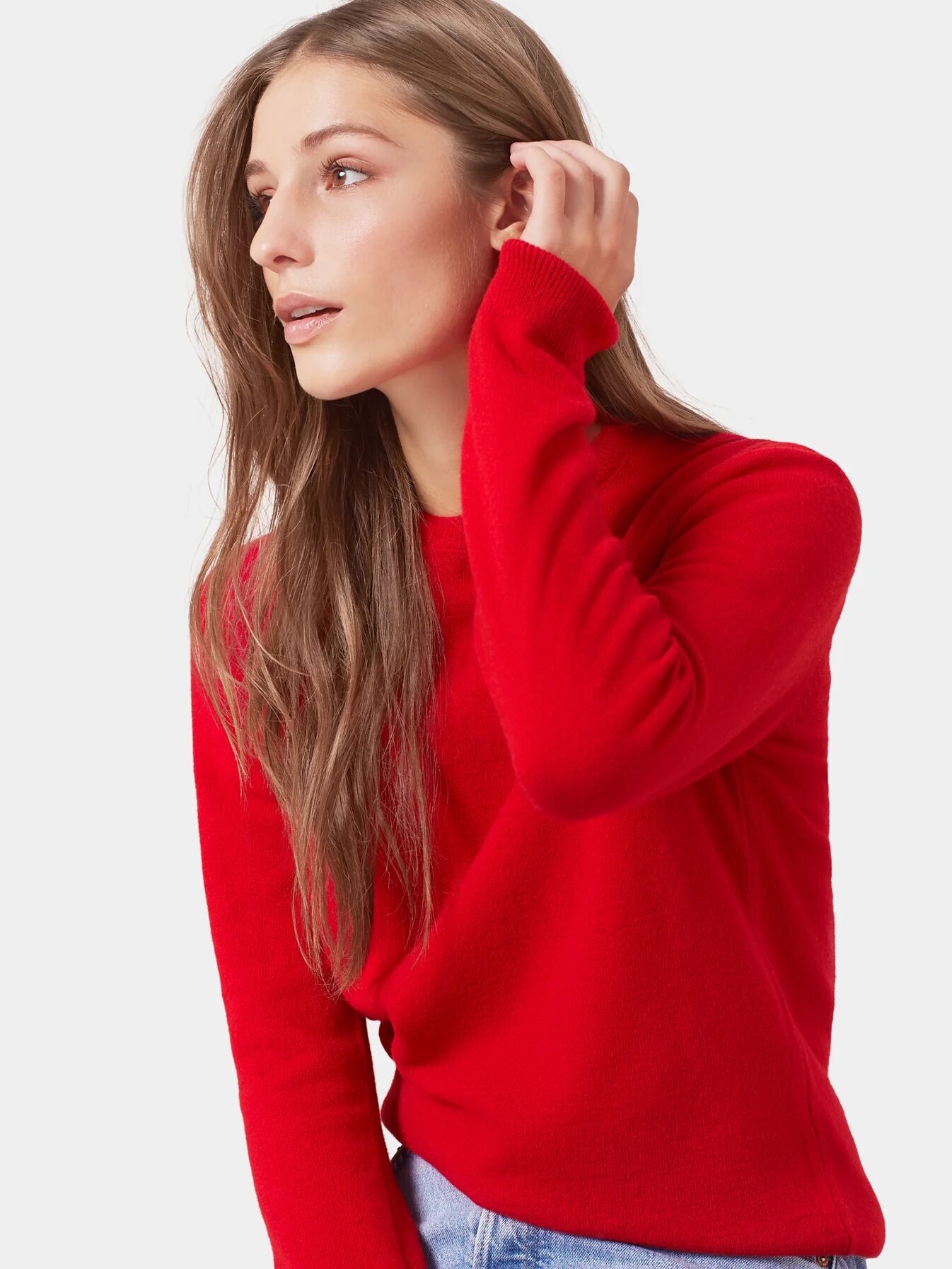 A woman with long brown hair wearing a bright red sweater and light blue jeans looks to the side with her hand near her face against a plain background.