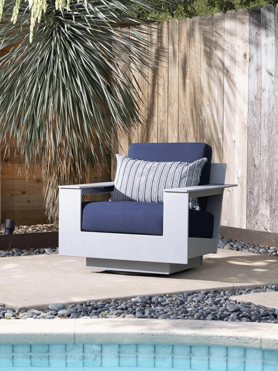 A modern outdoor chair with navy blue cushions and a striped pillow sits on a concrete patio near a wooden fence and a pool, surrounded by landscaping stones and plants.