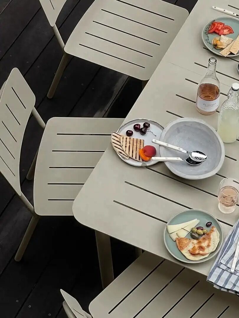 A beige outdoor table set with plates of fruit, crackers, cheese, drinks, and striped napkins, surrounded by matching chairs on a dark wooden deck.