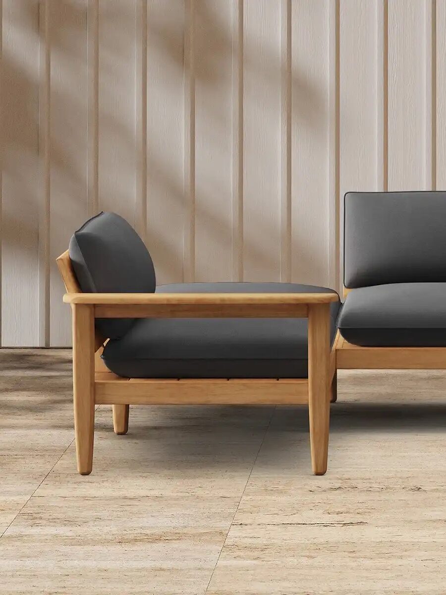 A wooden-framed outdoor sectional sofa with black cushions sits on a tiled patio against a vertical beige paneled wall.