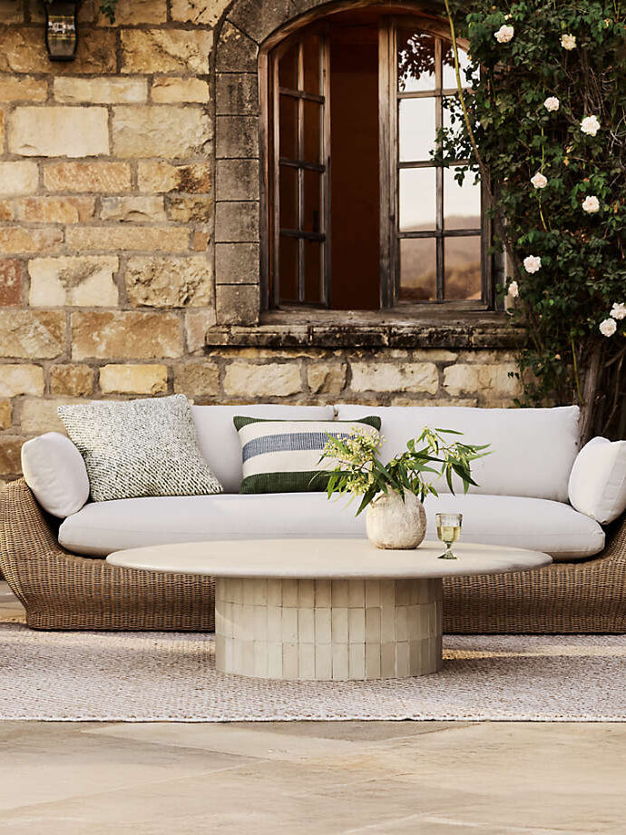 A wicker outdoor sofa with white and patterned cushions sits in front of a stone wall and window, next to a round table with a vase, glass, and greenery.