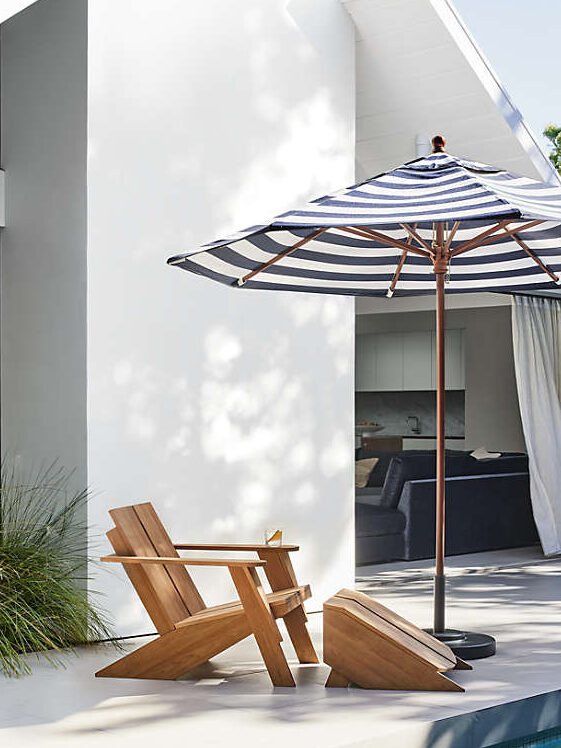 Two wooden lounge chairs with a matching footrest sit beside a swimming pool, shaded by a black-and-white striped patio umbrella near a modern white house.