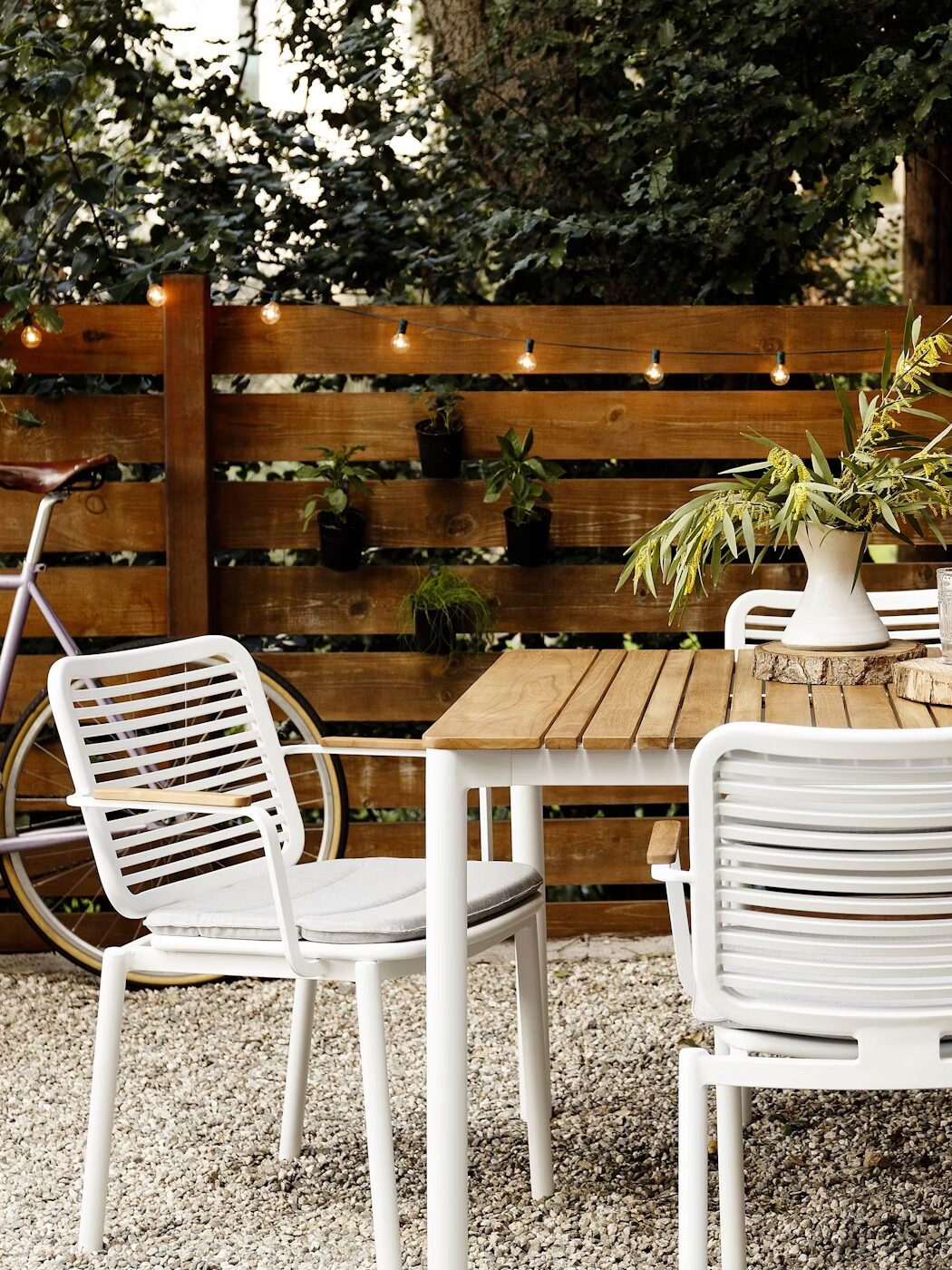 A wooden outdoor dining table with white chairs is set on gravel, decorated with plants and tableware, near a wooden fence with string lights and hanging plants.