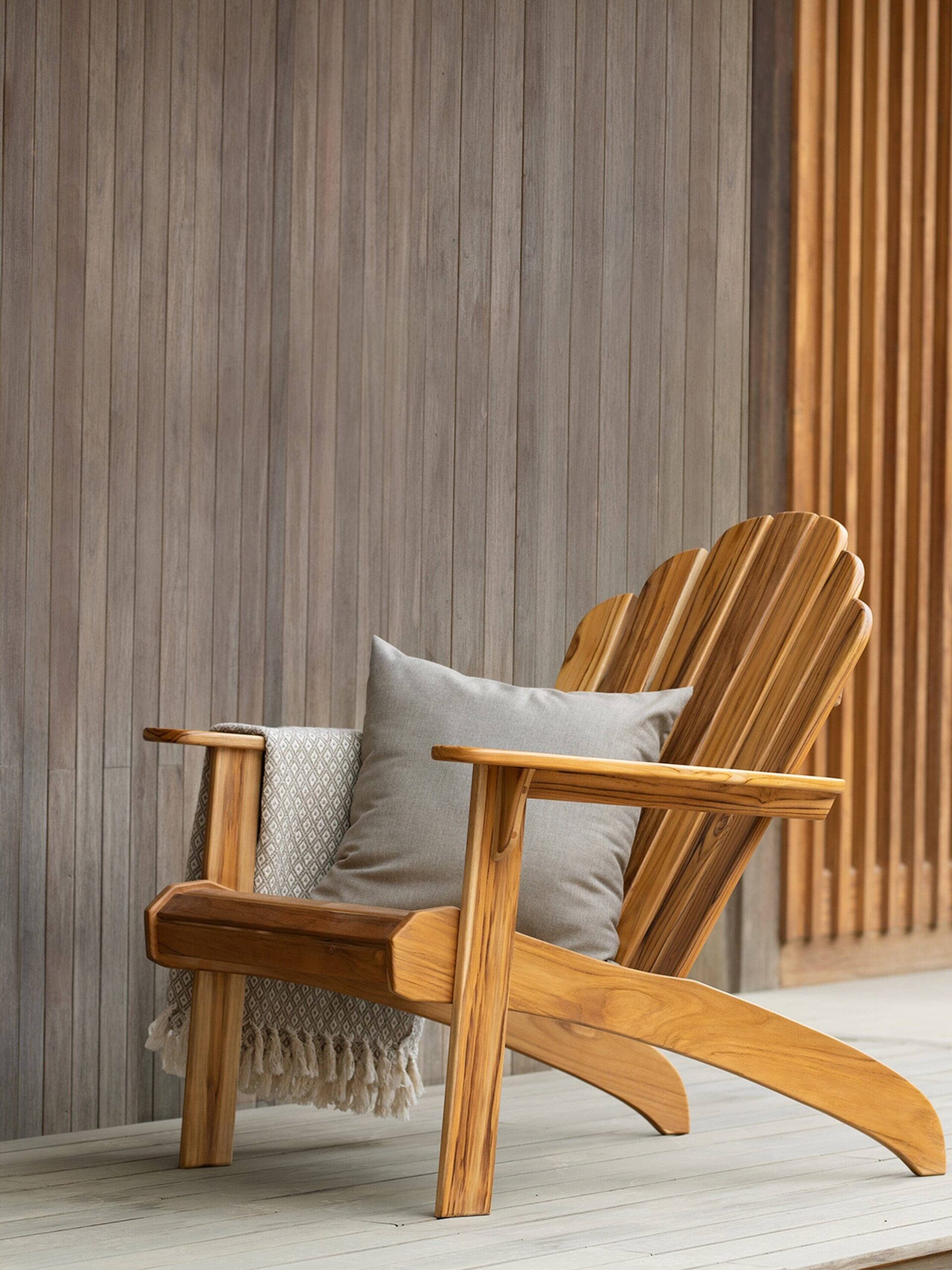 A wooden Adirondack chair with a beige cushion and a folded blanket sits on a wooden deck against a vertical slat wall.