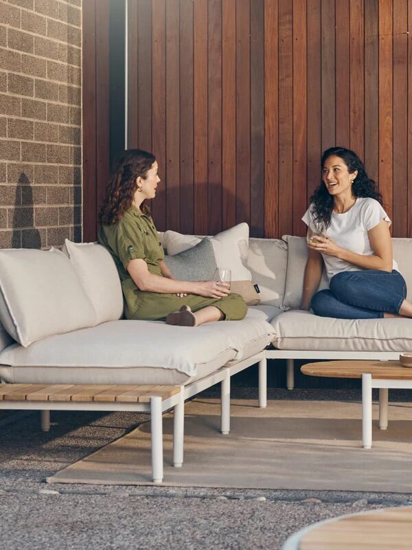 Two women sit and talk on a modern outdoor sectional sofa with drinks, next to a wooden coffee table with a wine bottle and snacks, against a wood and brick wall backdrop.
