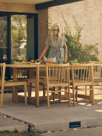 A woman with long gray hair stands by a wooden outdoor dining table set with drinks and dishes on a patio beside a modern house.