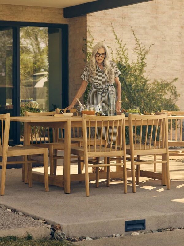 Two women set a wooden outdoor dining table with chairs on a patio beside a house with large windows and greenery.