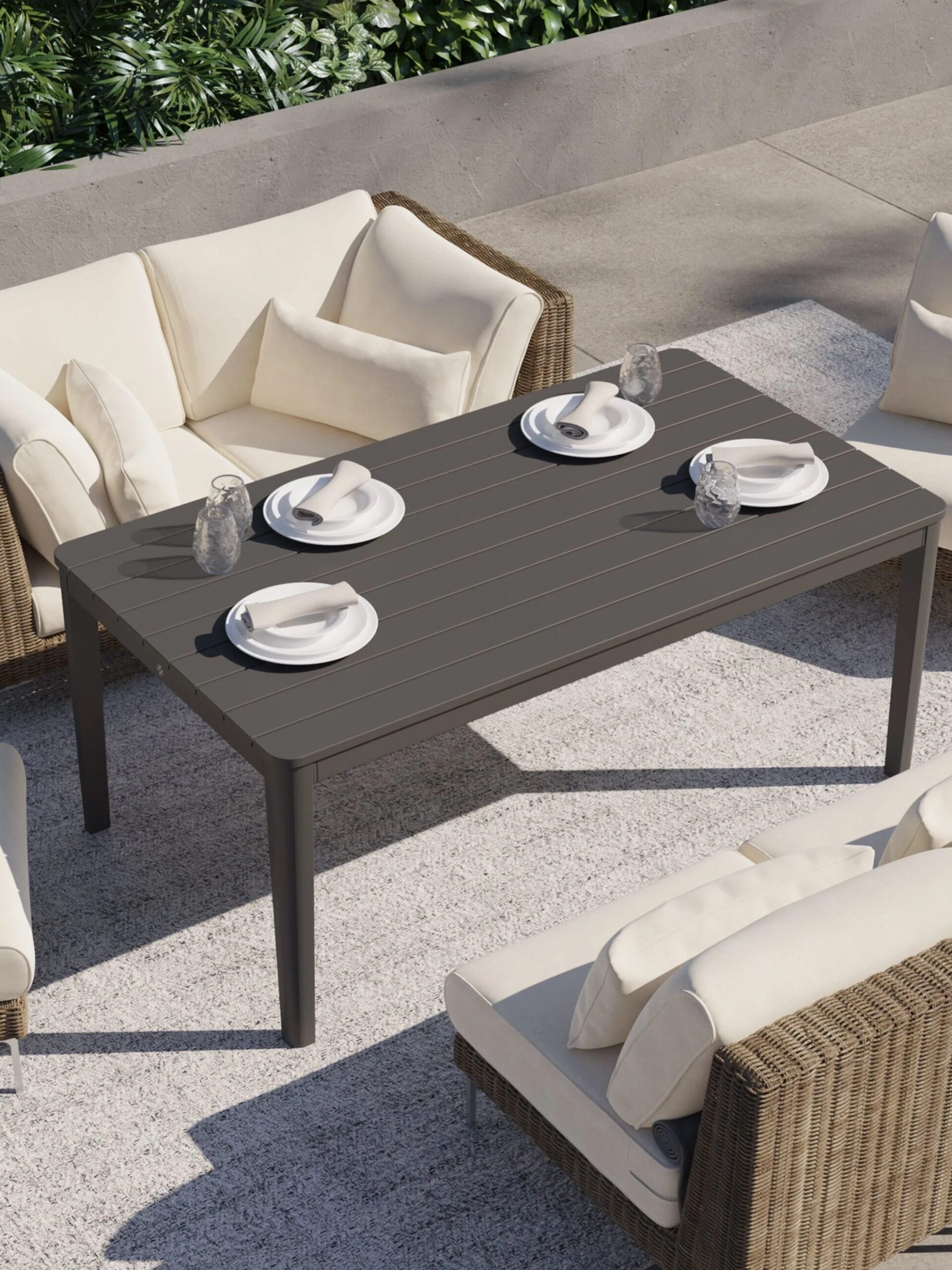Outdoor patio set with four cushioned wicker armchairs around a rectangular table set with four plates, napkins, and drinking glasses on a light-colored rug.