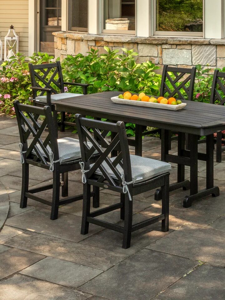 A black outdoor dining set with six cushioned chairs surrounds a rectangular table topped with a tray of oranges. The set is on a stone patio next to a house with green plants and flowers.