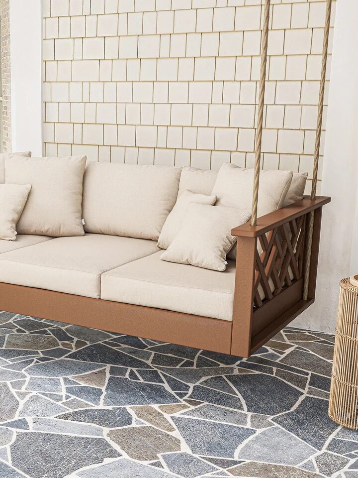 A wooden porch swing with beige cushions and pillows hangs on ropes above a stone tile patio, with a white tiled wall and greenery in the background.