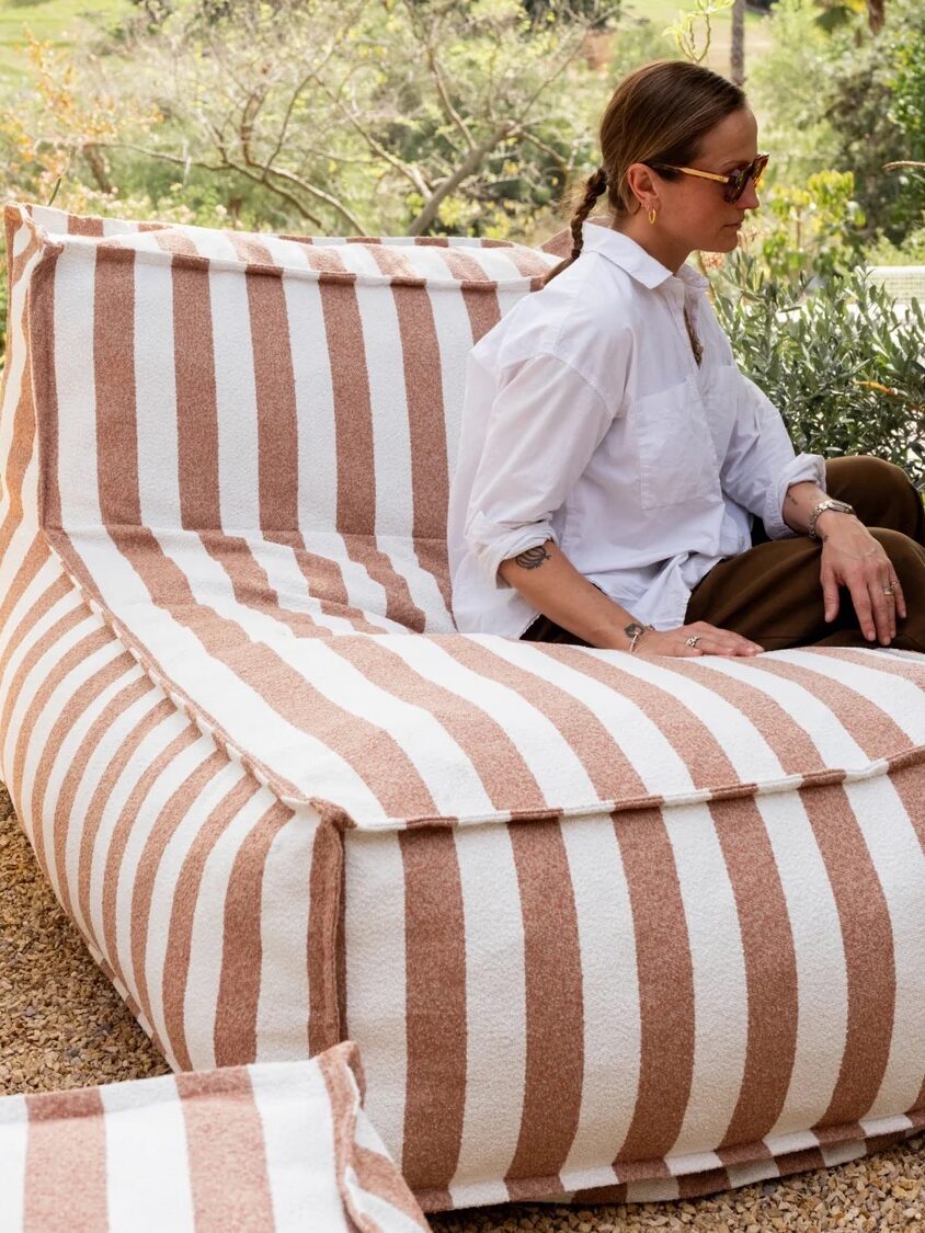A person wearing sunglasses and a white shirt sits on a large, brown-and-white striped outdoor lounge chair surrounded by plants and rocks.