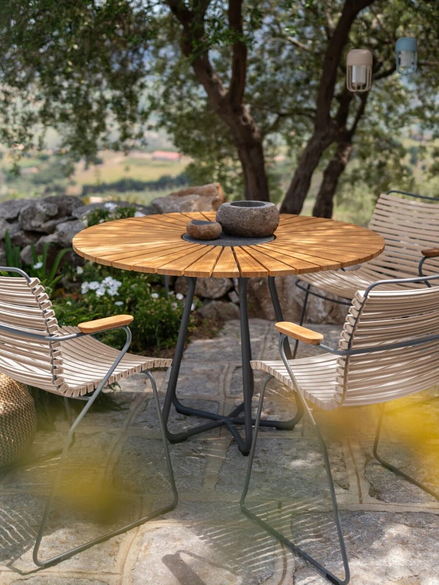 A round wooden table with three wicker chairs sits on a stone patio outdoors, surrounded by trees and greenery. Stone bowls are on the table.