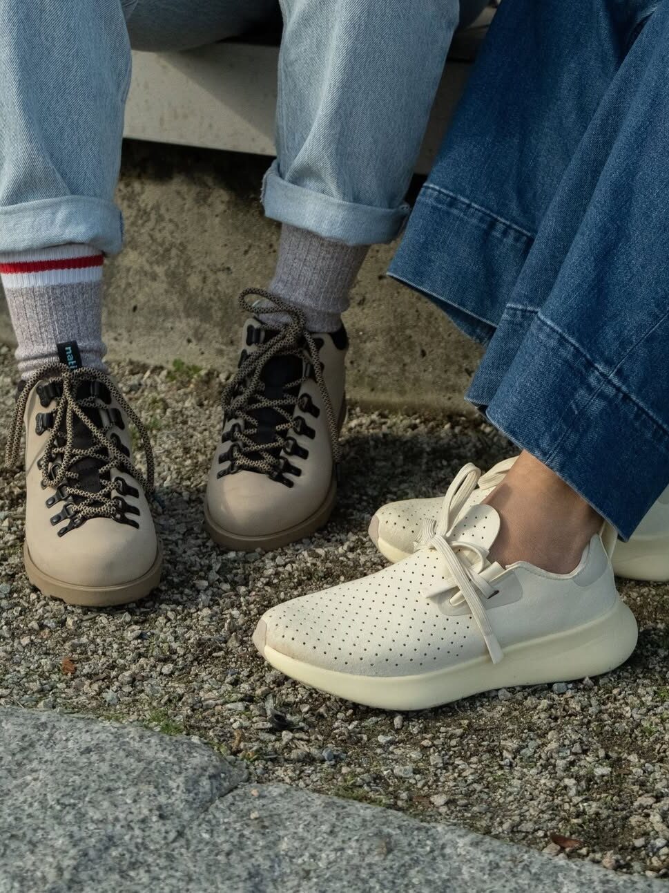 Two people wearing light-colored shoes—one in hiking boots and cuffed jeans, the other in sustainable sneakers and wide-leg denim—standing on a gravel path.