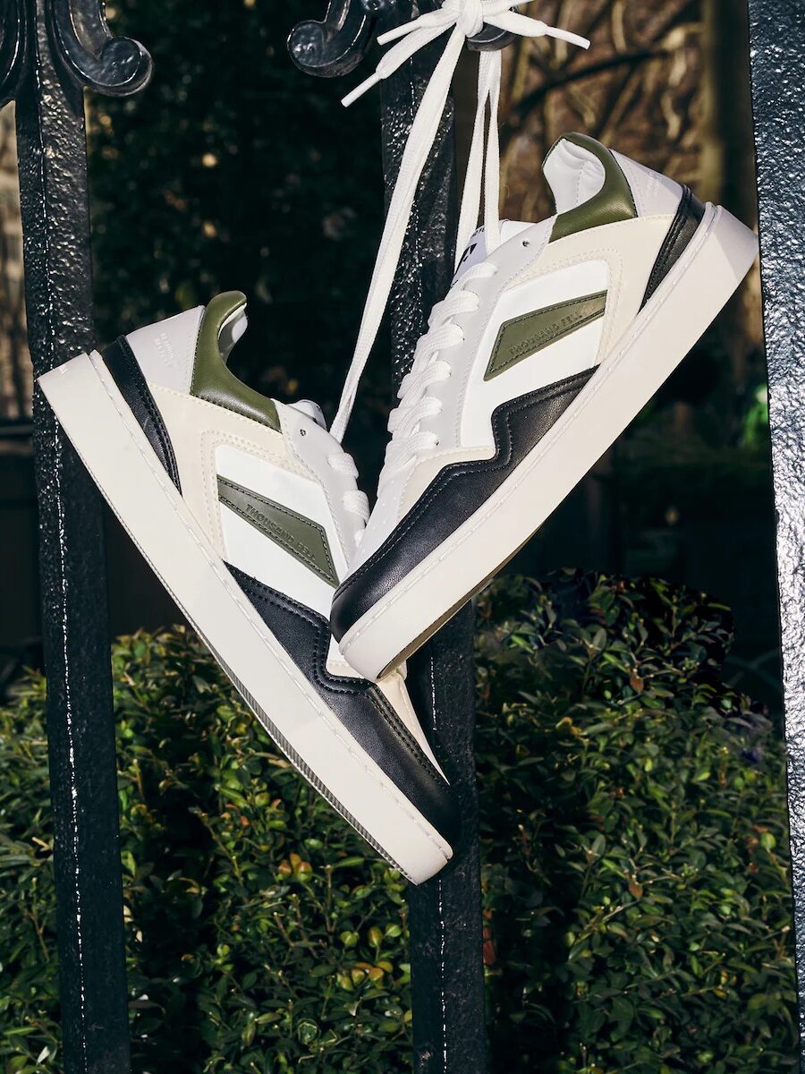A pair of white, green, and black sustainable sneakers hangs by their laces from a black iron fence, with greenery and a blurred outdoor background.