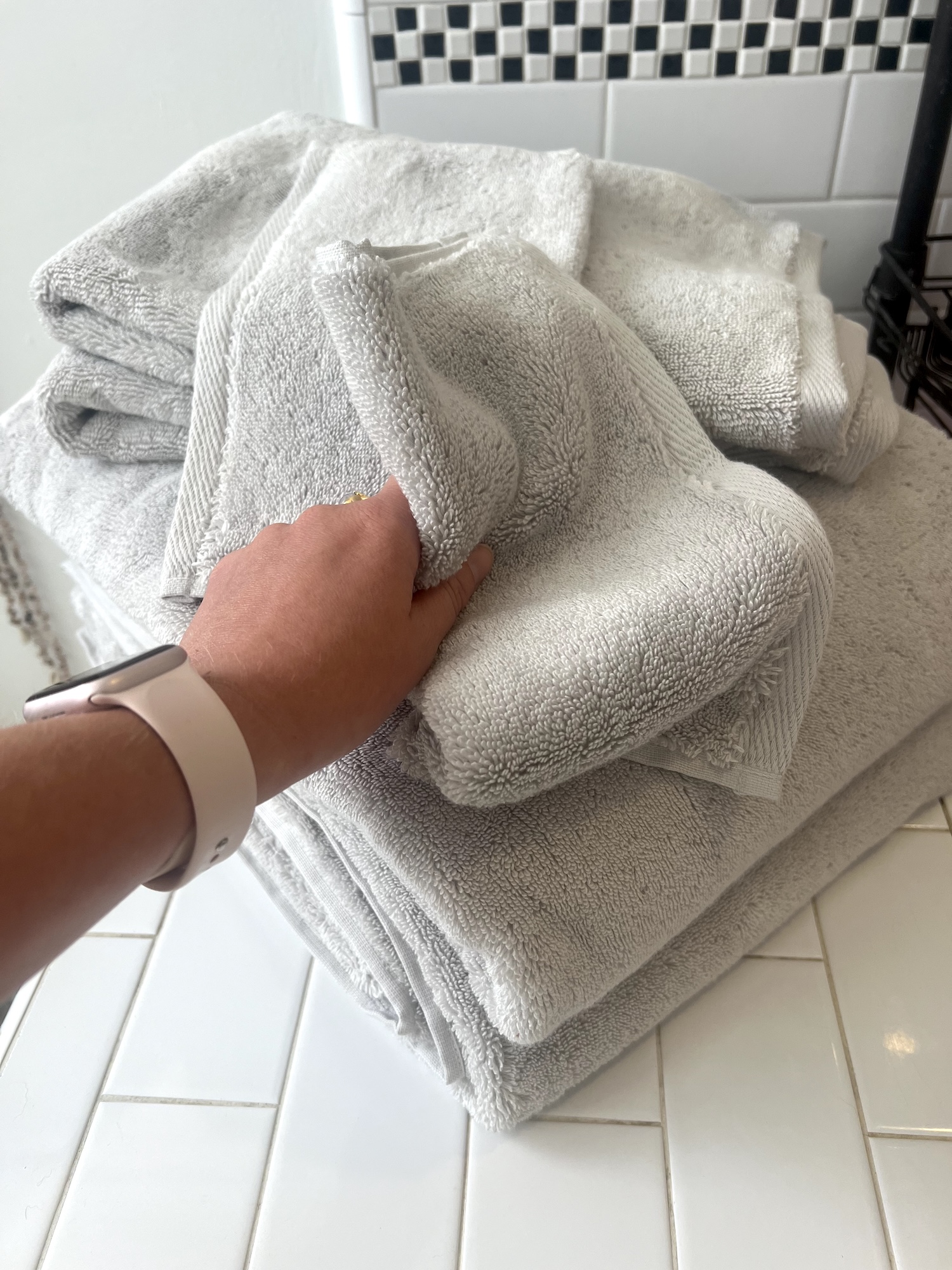 A person wearing a smartwatch grabs a folded gray organic towel from a stack of towels on a white tiled surface in a bathroom.