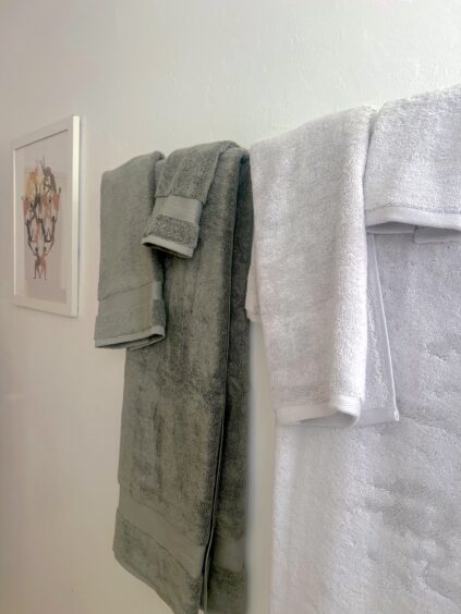 Two organic towels, one gray and one white, hang side by side on a rack against a white wall; a framed artwork is visible on the left side of the image.