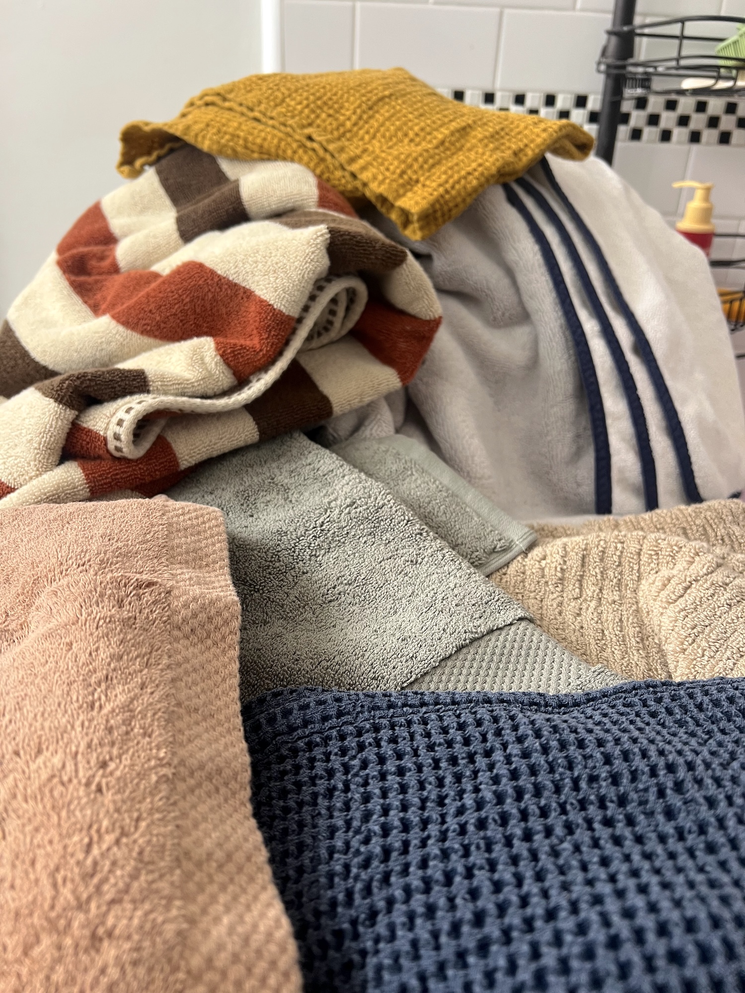A pile of assorted organic towels in various colors and textures is stacked in a bathroom, with toiletries and tiled walls visible in the background.