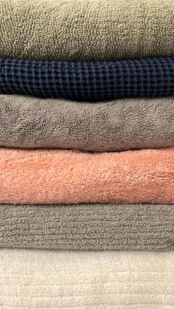 A stack of six folded organic towels in various colors and textures, including beige, navy, gray, and peach.