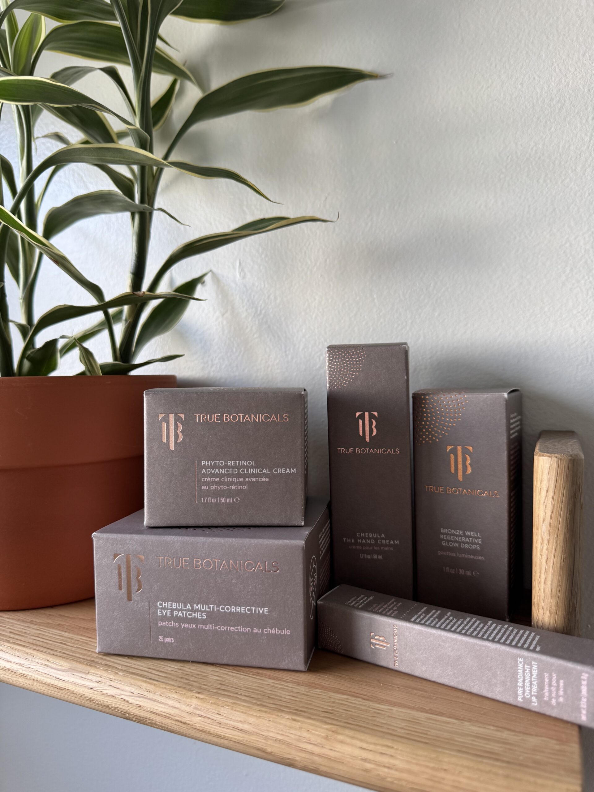 Boxes of True Botanicals skincare products are neatly arranged on a wooden shelf next to a potted plant against a light-colored wall.